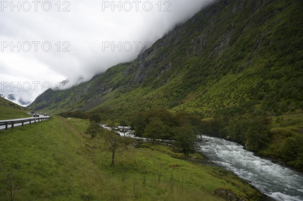 Byrkjelo, Vestland, Norway, Lonely road and river through a green valley surrounded by mist-covered mountains