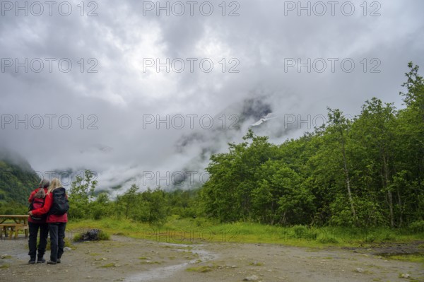 Fjærland, Vestland, Norway, couple looking at misty mountains surrounded by thick forest and a cloudy sky