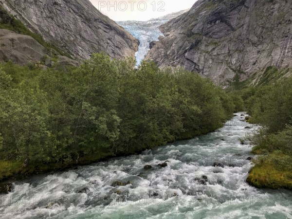 Briksdalsbre, Vestland, Norway, A rushing river flows through a green forest, with the Brikdalsbreen glacier and its trough valley in the background, framed by high mountains