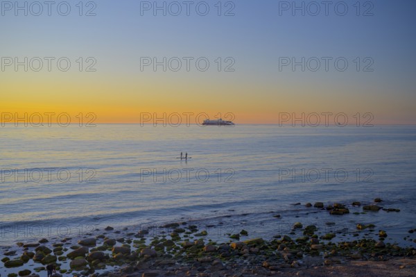 Hirtshals, Nordjylland region, Denmark, sunset over a calm sea with gentle waves and rocks on the shore in the background a ferry from Hirtshals to Kristiansand in the foreground two stand-up paddlers