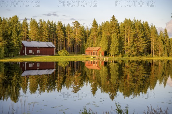Östanå, Torsby, Värmland, Sweden, Two red buildings by the lake, perfectly reflected in calm water, surrounded by a thick forest