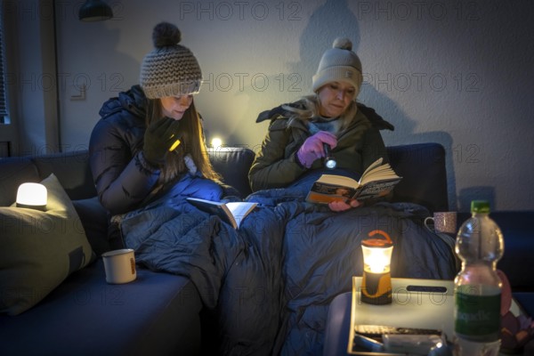 Symbolic picture blackout, extensive, prolonged power failure, in private surroundings, without electricity, heating, water, family on the sofa with warm winter clothes, sleeping bag, light from battery lights, battery lights, flashlights