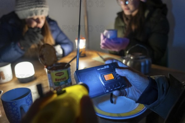 Symbolic blackout, extensive, prolonged power failure, in private surroundings, without electricity, heating, water, family at the dining table with warm winter clothes, sleeping bag, battery-operated radio, only source of information, no TV, Internet, mobile phone, light from battery lights, flashlights, food from cans, ready meals, heated on the camping stove, limited supply of food