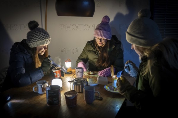 Symbolic blackout, extensive, prolonged power failure, in private surroundings, without electricity, heating, water, family at the dining table with warm winter clothes, sleeping bag, light from battery lights, flashlights, canned food, ready meals, heated on the camping stove, limited supply of food