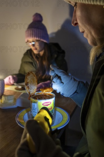 Symbolic blackout, extensive, prolonged power failure, in private surroundings, without electricity, heating, water, family at the dining table with warm winter clothes, sleeping bag, light from battery lights, flashlights, canned food, ready meals, heated on the camping stove, limited supply of food