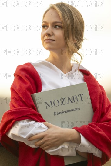 Self-confidently looking young woman, pianist, in white blouse and red cardigan, in the evening light, holding notes with Mozart, piano sonatas, in her arms, Stuttgart, Baden-Württemberg, Germany