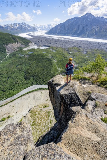 Young woman on a rocky outcrop enjoying the view, view of impressive mountain landscape with Matanuska glacier and glaciated mountain peaks, Lion's Head, Chugach Mountains, Alaska, USA