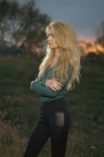 A woman stands with her arms crossed on a grassy hill. She faces the horizon where the sun is setting, creating a colorful sky. Trees and a vehicle are visible in the background