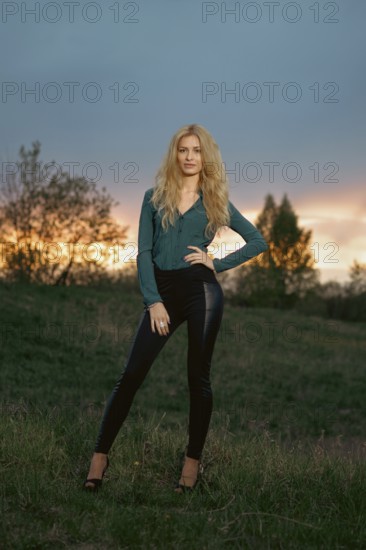 A woman poses on a grassy hill at sunset. The sky shows shades of orange and purple. Trees are in the background, and the setting appears to be natural and open