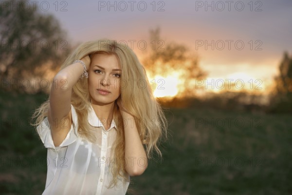 A woman stands in a field with blonde hair flowing as she poses during sunset. The sun creates a warm glow behind her, enhancing the nature around. She looks directly at the camera