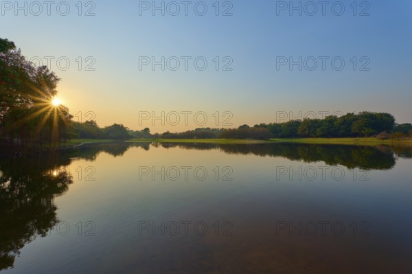 The first rays of sunshine fall on the calm lake, surrounded by green trees and clear skies, Fazenda Barranco Alto, Rio Negro, Pantanal, Mato Grosso, Brazil