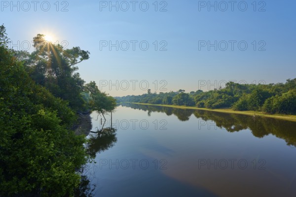 Sun rays shine through trees across the calm river, blue sky as backdrop, Fazenda Barranco Alto, Rio Negro, Pantanal, Mato Grosso, Brazil