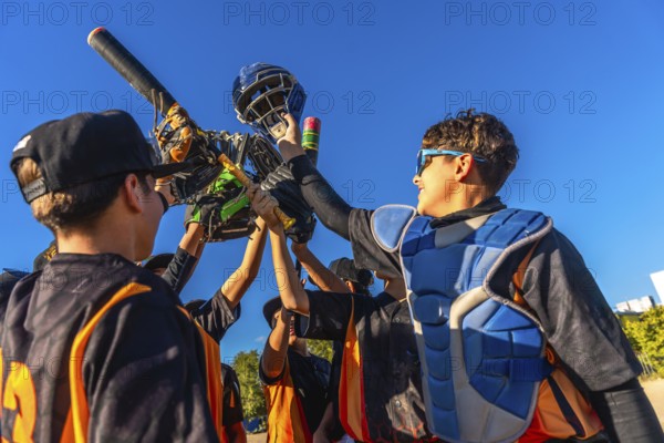 Young baseball players huddling together, lifting catcher's gear and a bat into the bright blue sky, celebrating a team achievement and showing unity on the field