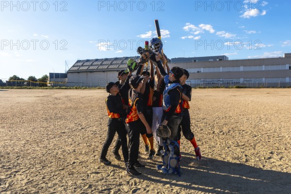 Young baseball players huddling together with arms raised in victory, holding bats, gloves, and helmets high celebrating their team accomplishment on a dirt field under a clear blue sky