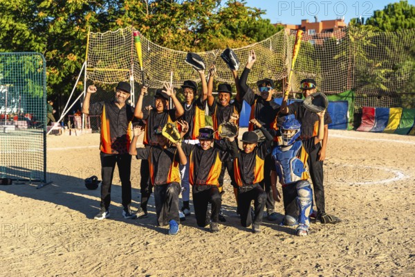 Young baseball team and coach standing together on an outdoor dirt field, raising bats and gloves in celebration of a game well played, representing sportsmanship and achievement