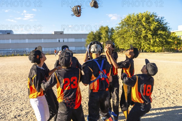 Young baseball players with gloves and uniforms celebrating a game victory together on a dirt field, looking up at two tossed gloves in the air under a clear blue sky