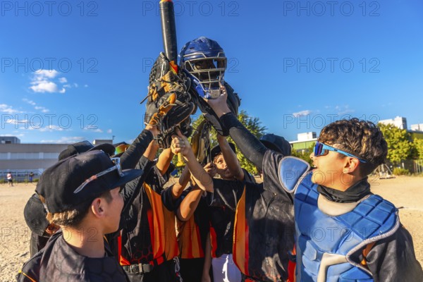 Baseball players huddling together, raising their catcher's helmet, bat, and gloves in the air, expressing team unity and triumph after a game on a sunny day