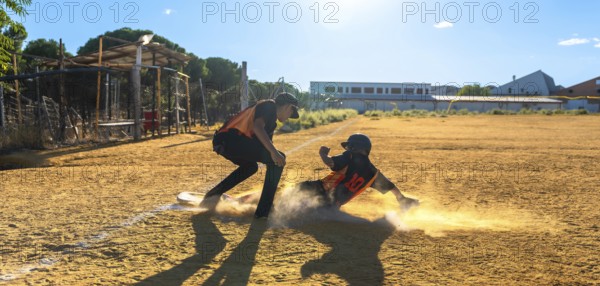 Two baseball players on a sunny field, one slides into a dusty base in a cloud of dirt while teammate watches the play, capturing speed, impact and competitive action