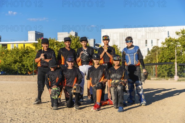 Young baseball players wearing uniforms and protective gear on a sunny day, standing and kneeling on a dirt field with trees and buildings in the background