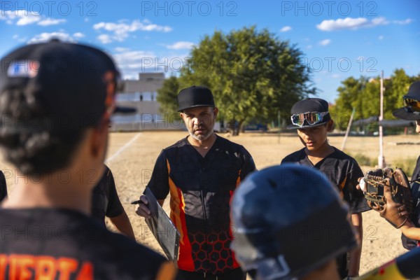 Baseball coach in a black and orange jersey and cap huddling with his youth team, discussing game strategy before or during a match on a dusty field under a clear sky