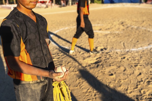 Young baseball boy in orange and black uniform holds glove and ball on sunlit dirt field during game, focused and ready while teammate stands blurred in background