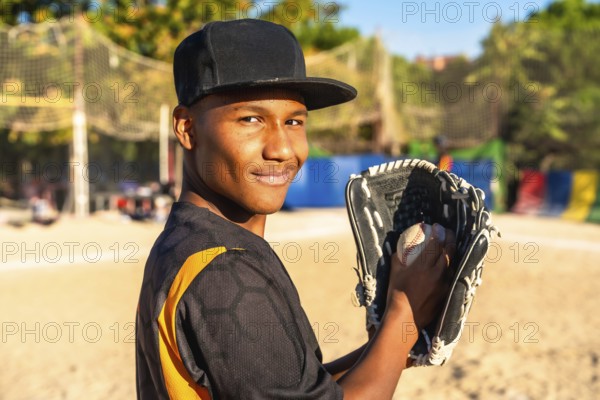 Young male baseball player stands on a sunny field, smiling confidently while holding a baseball in his glove, ready for practice or game with relaxed, focused energy