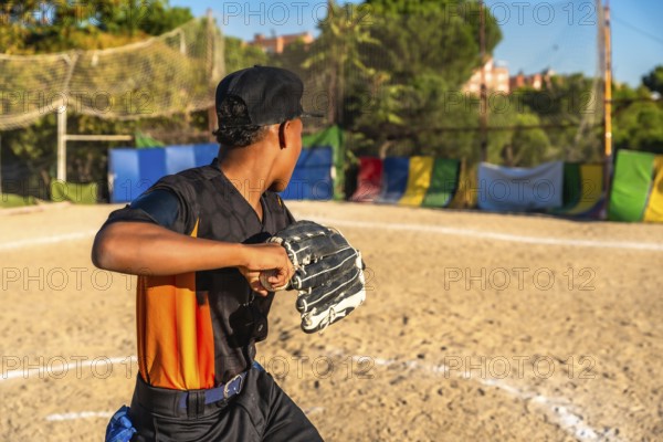 Young boy playing baseball, wearing a glove and cap, holding a ball, practicing pitching on a dirt field with colorful barriers in the background during a sunny outdoor game