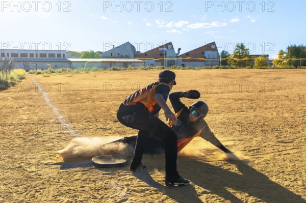 Baseball player sliding into the base with dust kicking up while an opponent stands by, capturing the intensity and action of a competitive game on a sunny day