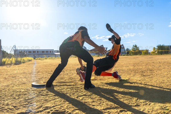 Two young baseball players are competing on a dusty field. With one player sliding dramatically toward base while the other is reaching out. Capturing the intense action of the game under a bright sky