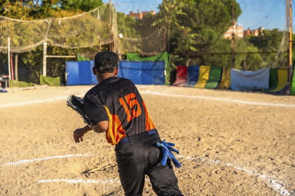 Player wearing a baseball uniform, cap, and fielding glove on a dirt field, throwing a ball during a game or practice session on a sunny day with a netting in the background