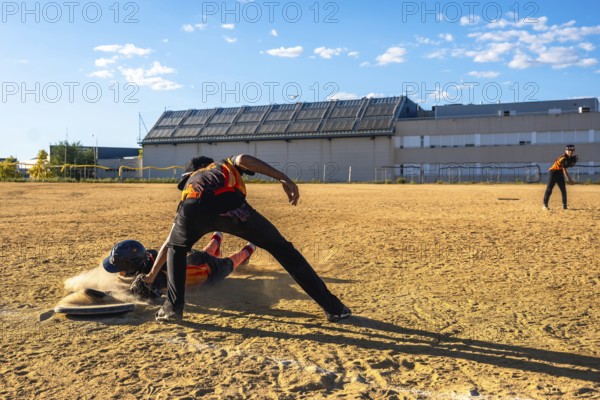 Baseball player sliding into home plate in a cloud of dust, attempting to score as the catcher prepares to tag, while another fielder stands in the background on a sunny day