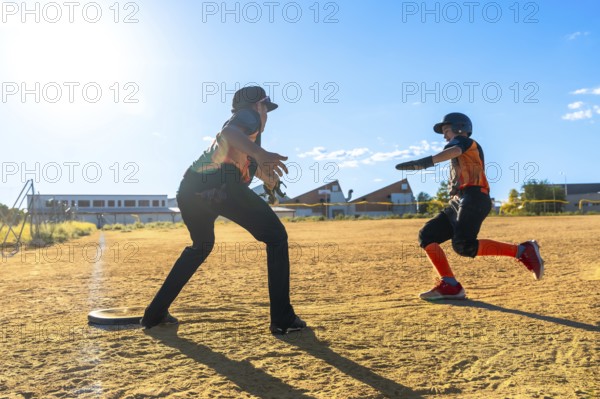 Youth baseball player sprinting hard across the dirt toward a base while coach or teammate awaits, showcasing speed, focus and summer youth sports action on a sunny field