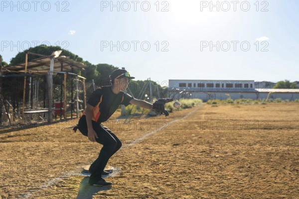 Baseball player in action on a dusty youth sports field, wearing uniform, cap and glove, extending his arm to catch a flying baseball under bright sunlight