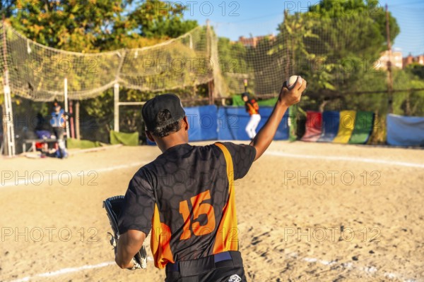 Young baseball pitcher in a black and orange uniform with number 15 on the back holding a ball ready to throw during a game on a sunny day at a dirt field