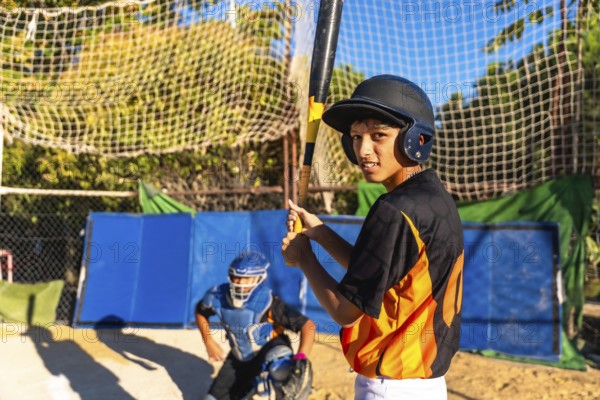Young baseball batter wearing a helmet and looking at the camera while holding a bat, with a catcher in full gear positioned behind him on the field