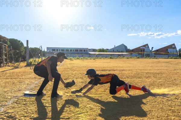 Kids playing baseball on a dusty field, one player sliding into home plate kicking up dust while another player waits ready with a glove, illustrating competition and youth sports