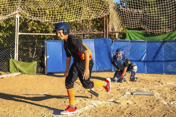 Young boy smiles while running on the dirt field, holding a bat and wearing a helmet and uniform, after hitting the ball during a youth baseball game as a catcher observes