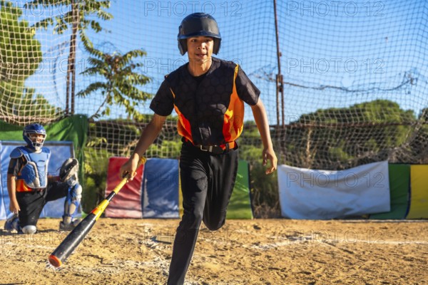 Young male baseball player running on a dirt field during a game or practice, wearing a helmet and uniform, holding a bat, with a catcher in the background
