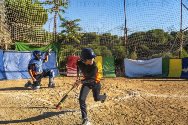 Kids playing baseball on a sunny day, an unidentifiable male batter wearing a helmet and looking back while running for first base and a catcher kneeling behind home plate