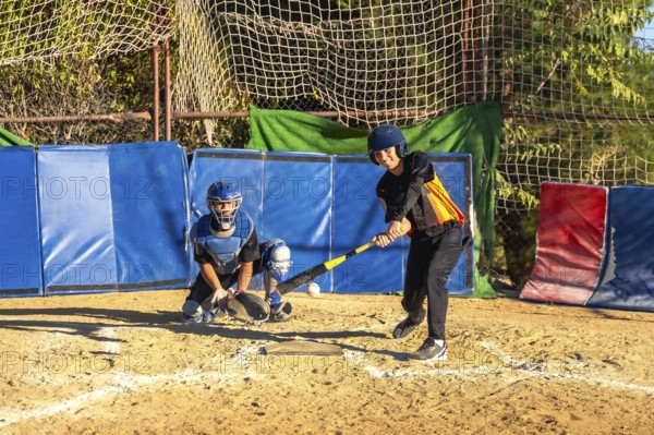 Youth baseball players, a batter taking a swing at a pitch and a catcher ready behind home plate, participating in a practice session on a dirt field with protective netting and padding