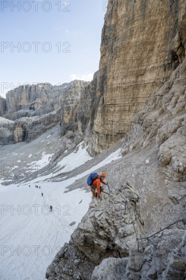 Mountaineer climbs on an exposed rock in the secured Via Ferrata Bocciere Centrale via ferrata, view of snow remains from the Vedretta del Sfulmini glacier, Brenta Mountains, Parco Naturale Brenta-Adamello, Trentino, Italy