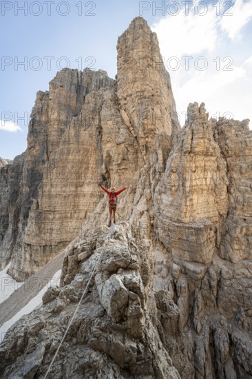 Mountaineer stretches his arms in the air, climbs an exposed rock in the secured Via Ferrata Bocciere Centrale via ferrata, mountain landscape with spectacular rock towers in the back, Brenta Mountains, Parco Naturale Brenta-Adamello, Trentino, Italy