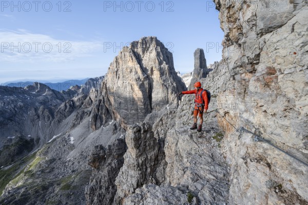 Mountaineer points into the distance, climbs an exposed rock band in the secured Via Ferrata Bocciere Centrale via ferrata, spectacular mountain landscape with steep rocky peaks, Brenta Mountains, Parco Naturale Brenta-Adamello, Trentino, Italy