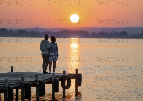 A couple stands on a jetty and looks at a colorful sunset over Lake Dümmer, Lembruch, Dümmer nature park Park, Lower Saxony, Germany