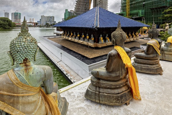 Buddha statues with yellow cloths sit in front of a modern cityscape on the water, The Seema Malaka Temple with the Buddha Statues in Colombo Sri Lanka
