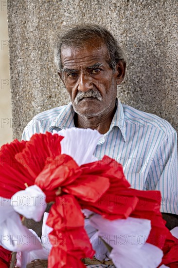 Close-up of an elderly man with red and white decorative flowers in the foreground, people making garlands in Colombo Sri Lanka City Hall