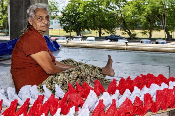 Elderly woman sitting outdoors working on red and white decorations, people making garlands in Colombo Sri Lanka City Hall