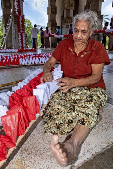 Elderly woman working with red and white decorations in an open hall, people making garlands in Colombo Sri Lanka City Hall