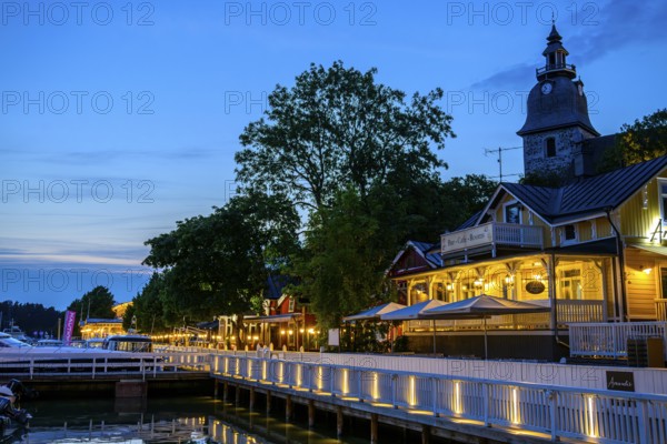 Street scene on Naantali harbour promenade, illuminated buildings and waterside boats at dusk, with church in the background, Naanatali, Varsinais-Suomi, Finland