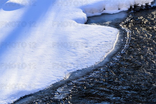 Ilsenburg, Saxony-Anhalt, Germany, A partially frozen Ilse river in the Harz National Park on Heinrich-Heine Weg Ilsetal hiking trail in winter with snow-covered banks and clear water, a clearly marked ice edge with blue snow in front of flowing water